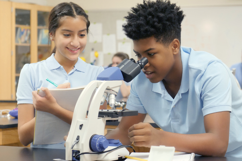 One student looking into a microscope while another student observes.