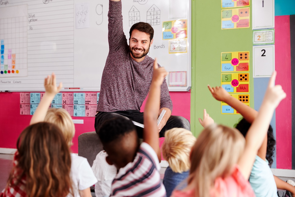 A teacher in front of a classroom full of students, everyone has their hand raised.